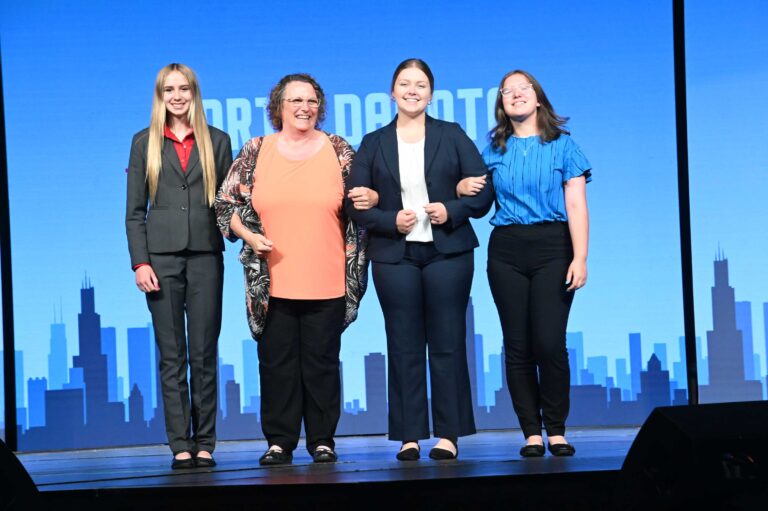Four people stand smiling on stage in front of a blue backdrop with a city skyline graphic, representing Youth Leadership Programs. Three young women in business attire stand beside an older woman in a patterned cardigan and orange shirt.