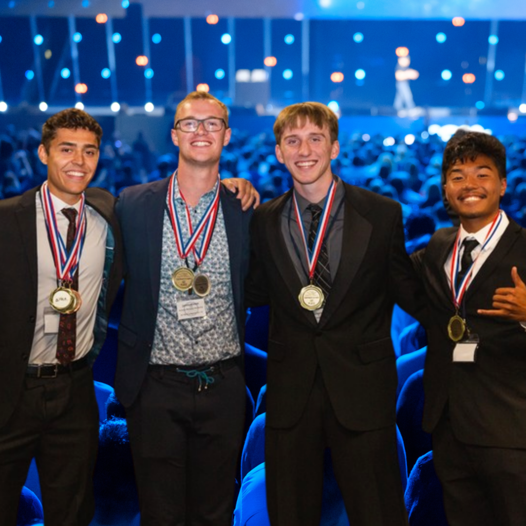 Four young men in suits, wearing medals and smiling, stand together with arms around each other. They are on a stage in front of an audience, blue lighting and a speaker in the background, celebrating their achievements through Business Education Programs.