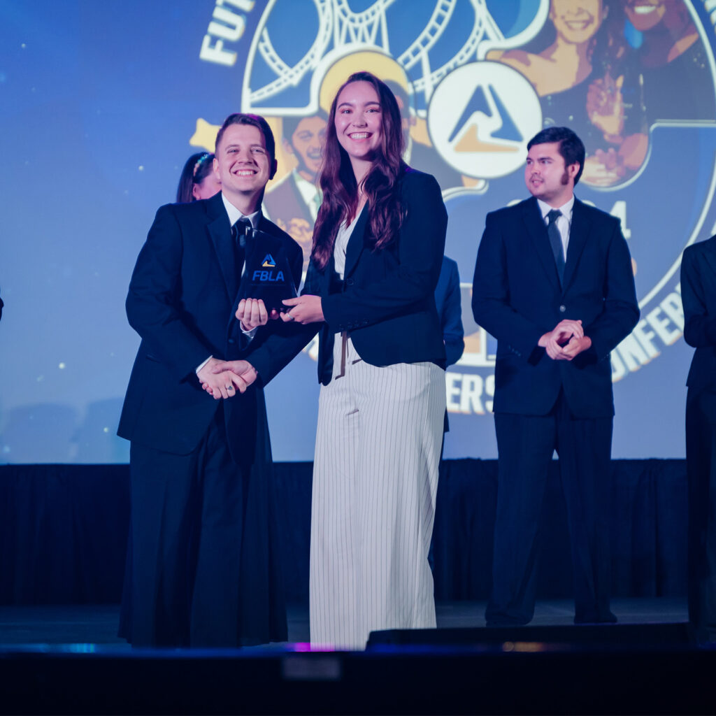 Two people in business attire shake hands and smile on stage while one holds an FBLA award, representing Business Education Programs. Other formally dressed individuals stand in the background. A large FBLA conference logo is projected behind them.