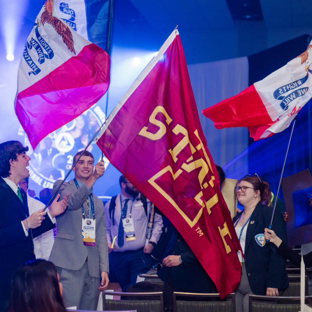 People in formal attire enthusiastically wave large flags, including a maroon one with yellow text and red, white, and blue ones, at an indoor event showcasing Business Education Programs amid bright stage lights and a lively audience.