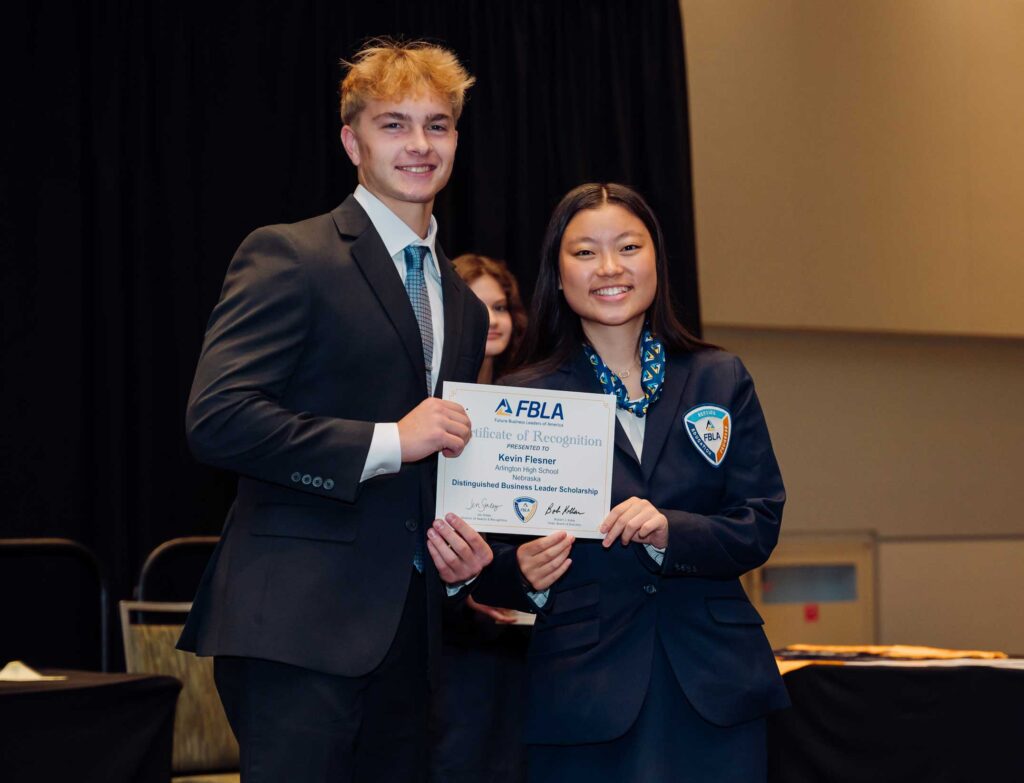 A young man in a suit stands next to a young woman in a blazer holding an Awards and Recognition certificate. Both are smiling and posing for the photo at an indoor event.