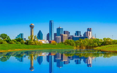 The Dallas skyline is reflected in a calm body of water, with a bright blue sky above and green grass and trees in the foreground. Iconic buildings, such as Reunion Tower, create a striking urban landscape.