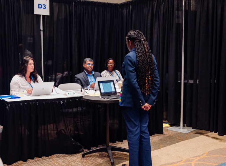 A person in a blue suit stands before a panel of three people seated at a table with laptops, delivering a presentation or participating in an interview during a professional conference.