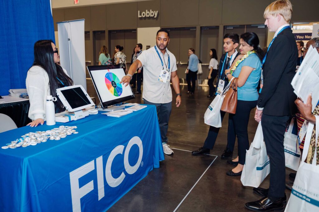 A man explains a colorful spinning wheel game at a FICO booth to a group of people at an indoor conference. A woman sits behind the table with FICO branding and promotional materials, engaging attendees in the activity.
