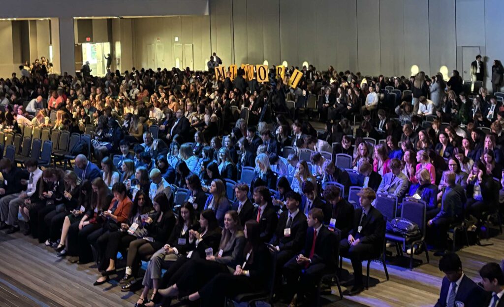 A large audience sits in rows of chairs at a conference inside a spacious hall. In the middle, people hold up signs that spell out MITFORD, creating a lively and professional atmosphere.