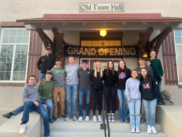 A group of sixteen smiling teenagers stands on steps outside Old Town Hall under a GRAND OPENING sign. It is evening, and the casually dressed group radiates excitement for the new chapter in their community.