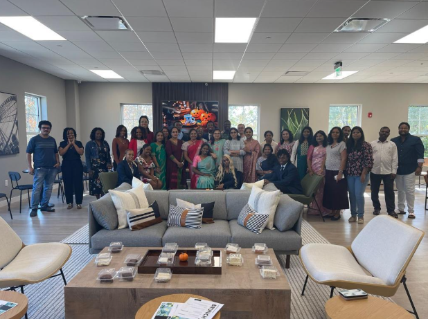 A group of people stands together in a spacious, well-lit room with modern decor, sofas, and chairs. Some are seated while most stand, all smiling and posing for a group photo beneath colorful artwork on the back wall.