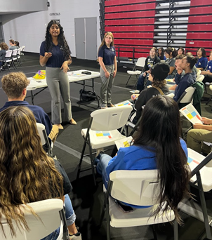 Two women stand at the front of a classroom speaking to a group of seated students, who are holding papers. The setting appears to be a gym with red bleachers in the background.