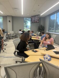 People sitting in groups at tables in a modern, well-lit meeting room with large windows. Some are talking and taking notes. There are laptops, water bottles, and bags on the tables. A presentation screen is visible in the background.