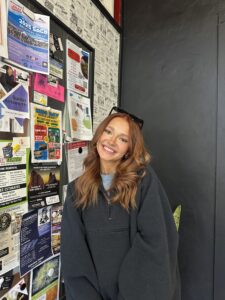 A woman with long, wavy red hair wearing a black jacket smiles while standing in front of a wall covered with colorful flyers and posters.