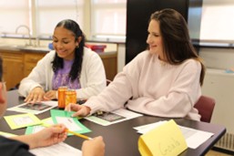 Two women sit at a table in a classroom, smiling and working with papers and colorful cards. One has dark, curly hair; the other has straight, light hair. There are name cards and a drink can on the table.