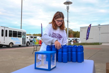 A young person wearing glasses and a Walk to End shirt arranges blue water bottles on a table outdoors, with a donation box and buses visible in the background.