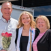 Three people stand smiling together outdoors; the man on the left holds a bouquet of red roses, the young woman in the middle wears a sash—perhaps celebrating at her daycare—and the woman on the right wears a striped top. A white truck and glass building are in the background.