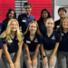 A group of thirteen smiling students wearing matching navy blue FBLA and DECA shirts with khaki pants pose together indoors in front of red bleachers and white tables.