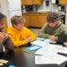 Five students sit around a table in a classroom, engaged in a financial literacy discussion. Papers, pencils, and some play money are spread out before them. Wooden cabinets and kitchen equipment can be seen in the background.