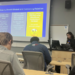A woman stands at the front of a classroom giving a presentation to seated students. Two large screens display slides about growth mindset and overcoming rejection, with text and emoji graphics visible.