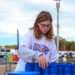 A young girl wearing glasses and a Walk to End event shirt arranges large blue blocks outdoors at an FBLA gathering, with buses and buildings visible in the background.