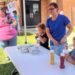 A man in a hat talks to a woman and three children at an outdoor table with wrapped food, drinks, and condiments. Other people are in the background near a brick building on a sunny day.