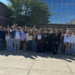 A large group of high school students, many showcasing leadership qualities, stand together outside a school building on a sunny day, posing for a group photo. Trees and building windows are visible in the background.