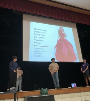 Four people stand on a stage in front of a large screen displaying a presentation titled How Owning Barbies Can Make You a Successful Business Person with an image of a Barbie doll in a pink dress.