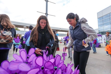 Two young women select large purple flower pinwheels at an outdoor event, surrounded by other participants wearing purple shirts. Buildings and a covered walkway are visible in the background.