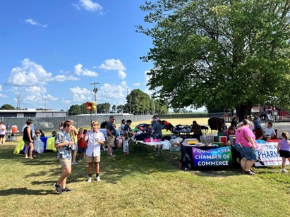 People gather outdoors at tables with banners, including one for the Chamber of Commerce, at a community event on a sunny day. Some people are walking or standing, while others browse items on tables under a large tree.