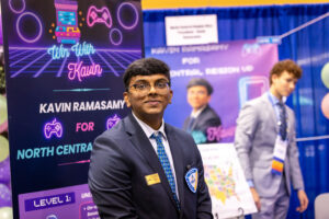 A young man in a suit and tie stands smiling in front of a colorful booth with gaming graphics and signs that read “Win With Kavin” and “Kavin Ramasamy for North Central.” Another person stands in the background.