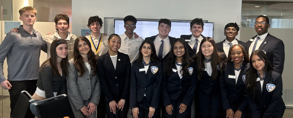 A group of sixteen young people, some in business attire and others in casual clothing, stand together and smile for a group photo in a modern office setting.