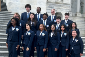 A group of thirteen young adults in matching navy suits with badges pose together on the steps of a marble building, smiling for a group photo.