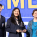 Three people stand on stage; a young man and a young woman in suits, with the woman holding an FBLA award, and an older woman in a blue outfit. A large blue and yellow backdrop is behind them.