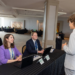 A young man in a light suit stands and speaks to two people sitting at a table with laptops in a conference room. The table has nameplates, and the setting appears to be a professional event or registration area.