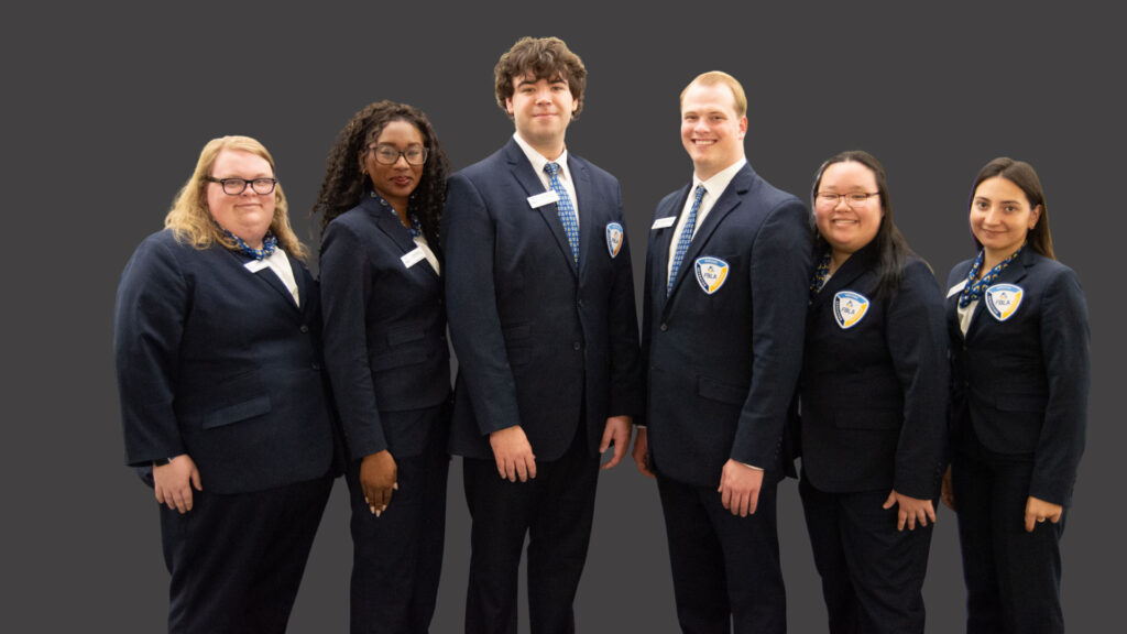 Six people wearing matching navy blue suits with name tags and badges stand side by side against a plain gray background, smiling and facing the camera.