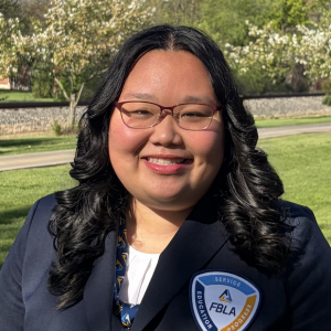 A person with long, wavy black hair and glasses smiles outdoors, wearing a dark blazer with an FBLA badge and a patterned tie. Trees and greenery are visible in the sunny background.