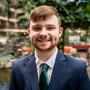 A young man with short brown hair and a beard, wearing a navy suit, white shirt, and green tie, smiles while standing indoors with greenery and blurred lights in the background.