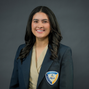 A woman with long dark hair and a beige top smiles while wearing a dark blazer with an FBLA emblem, standing against a plain dark background.