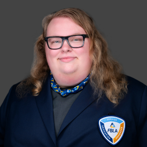 A person with long, wavy hair and glasses is wearing a dark blazer with an FBLA badge and a patterned scarf, smiling at the camera against a plain gray background.