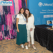 Two women smiling and posing together at a UWorld Accounting & Finance event booth, with colorful pink and purple streamers in the background and a table displaying branded items and snacks.