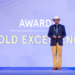 A man in a suit and white hat stands on stage holding an award, with the large text AWARD GOLD EXCELLENCE displayed on a blue-lit screen behind him.