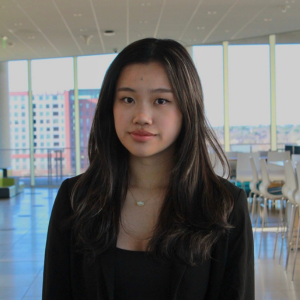 A young woman with long dark hair, wearing a black top and blazer, stands indoors in a bright, modern room with large windows and city buildings visible in the background.
