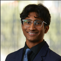 A young man with medium-dark skin, wavy dark hair, and glasses smiles at the camera. He is wearing a dark blazer, black shirt, and blue patterned tie, with a blurred background behind him.