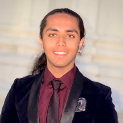 A young person with long dark hair pulled back, wearing a dark velvet suit jacket, a burgundy shirt, and a black tie, smiles at the camera against a blurred light-colored background.