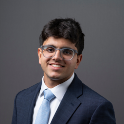 A young man with short dark hair and glasses, wearing a navy blue suit, white shirt, and light blue tie, smiles in front of a plain gray background.