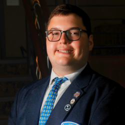 A man wearing glasses, a dark suit jacket with multiple pins, a blue patterned tie, and a white shirt smiles while standing indoors in front of a staircase.