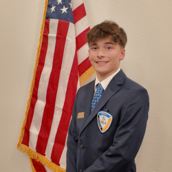 A young man in a blue suit stands smiling in front of an American flag. He wears a blue patterned tie and a badge on his jacket, suggesting participation in an official or student leadership event.