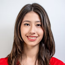 A young woman with long brown hair and brown eyes smiles at the camera. She is wearing a red top and a necklace, with a plain light-colored background behind her.