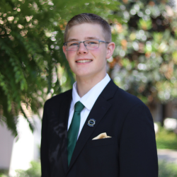A young man wearing glasses, a dark suit, white shirt, and green tie stands outside in front of leafy greenery, smiling at the camera.