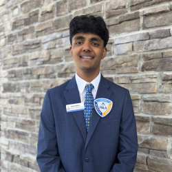 A young man in a blue suit and patterned tie smiles while standing in front of a stone wall. He wears a badge and a DECA pin on his jacket.