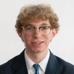 A young person with curly blond hair and glasses is wearing a dark suit, white shirt, and a blue patterned tie, smiling at the camera in front of a plain white background.