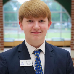 A young man with light brown hair in a navy suit, patterned tie, and white shirt, wearing a name badge that says “Grant Horton.” He is standing indoors with large windows and greenery in the background.
