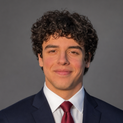 A young man with curly brown hair, wearing a dark suit, white shirt, and red tie, smiles slightly in front of a plain gray background.